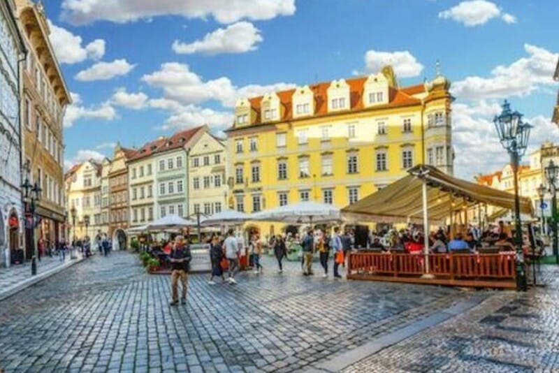 푸르고 흐린 하늘 아래 화려한 유서 깊은 건물로 둘러싸인 야외 카페에서 사람들이 산책하고 식사하는 자갈 광장.