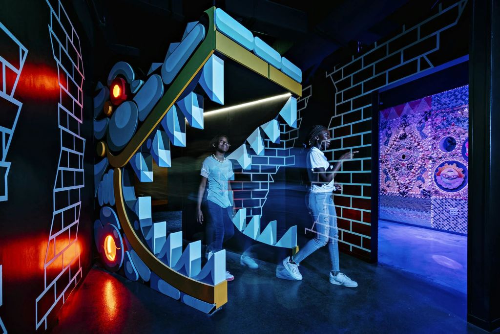 조명이 희미하고 다채로운 전시 공간에서 두 사람이 커다랗고 만화 같은 입이 열린 드래곤 디스플레이를 거닐고 있습니다.