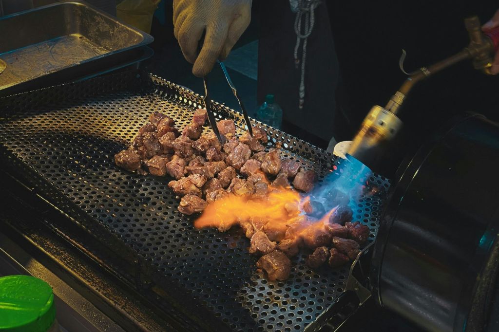 한 사람이 구멍이 뚫린 그릴에 집게와 눈에 띄는 불꽃이 있는 송풍구를 사용하여 고기 덩어리를 시어즈합니다.