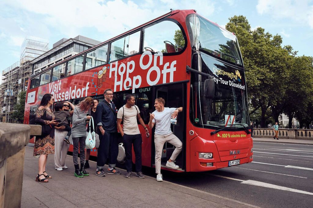 빨간색 2층 관광 버스 근처에 서 있는 한 무리의 사람들. "Hop On Hop Off" 가 적혀 있습니다.