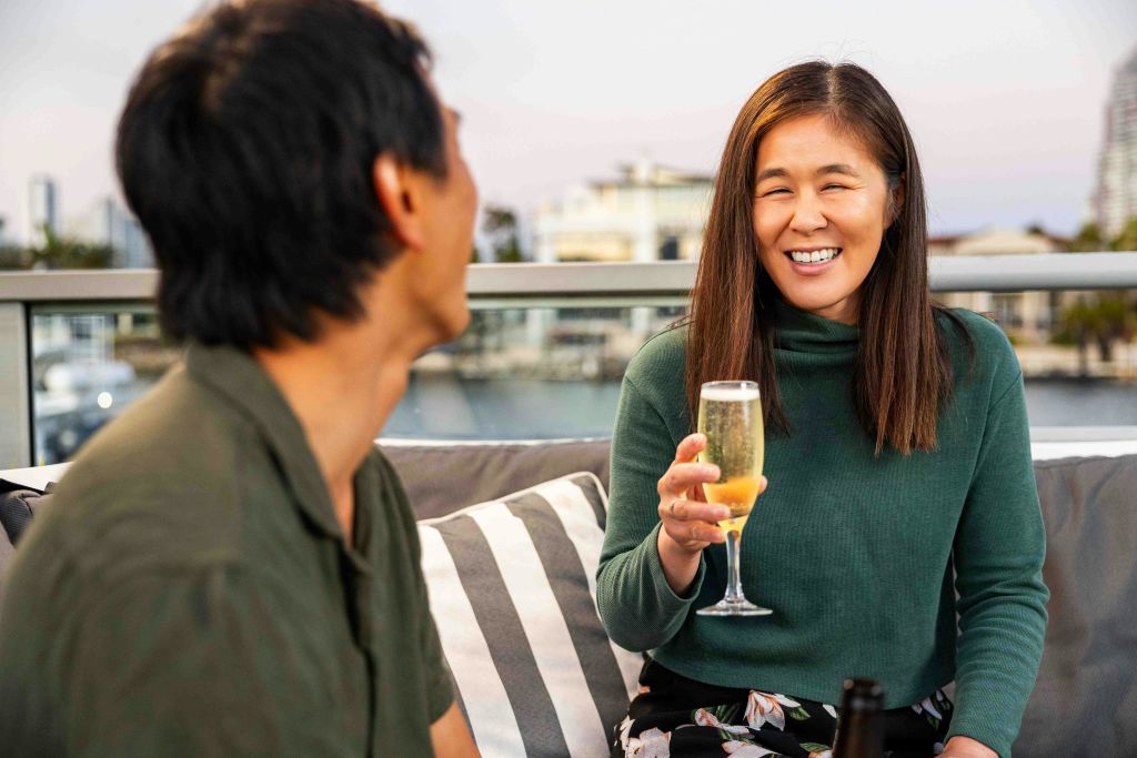 야외에서 줄무늬 쿠션이 놓인 소파에 앉아 웃으며 샴페인 한 잔을 들고 있는 여성.