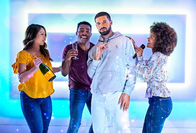 네온 조명이 켜진 방에서 세 명의 여성과 한 명의 남성이 음료와 마이크를 들고 즐거운 포즈를 취하고 있습니다.