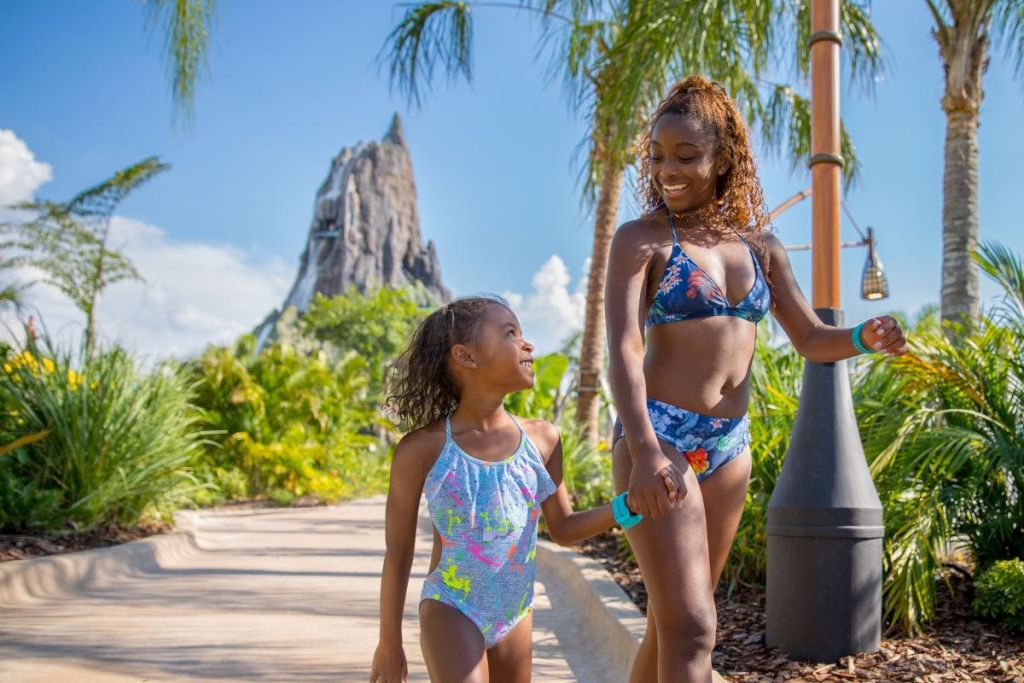 수영복을 입은 미소 짓는 두 소녀가 야자수와 산을 배경으로 열대 지방의 길을 함께 걷고 있습니다.