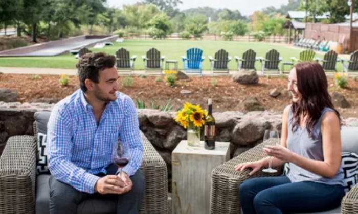 한 남성이 야외에서 고리버들 의자에 앉아 와인잔을 들고 있습니다. 배경에는 애디론댁 의자와 울창한 정원이 있습니다.