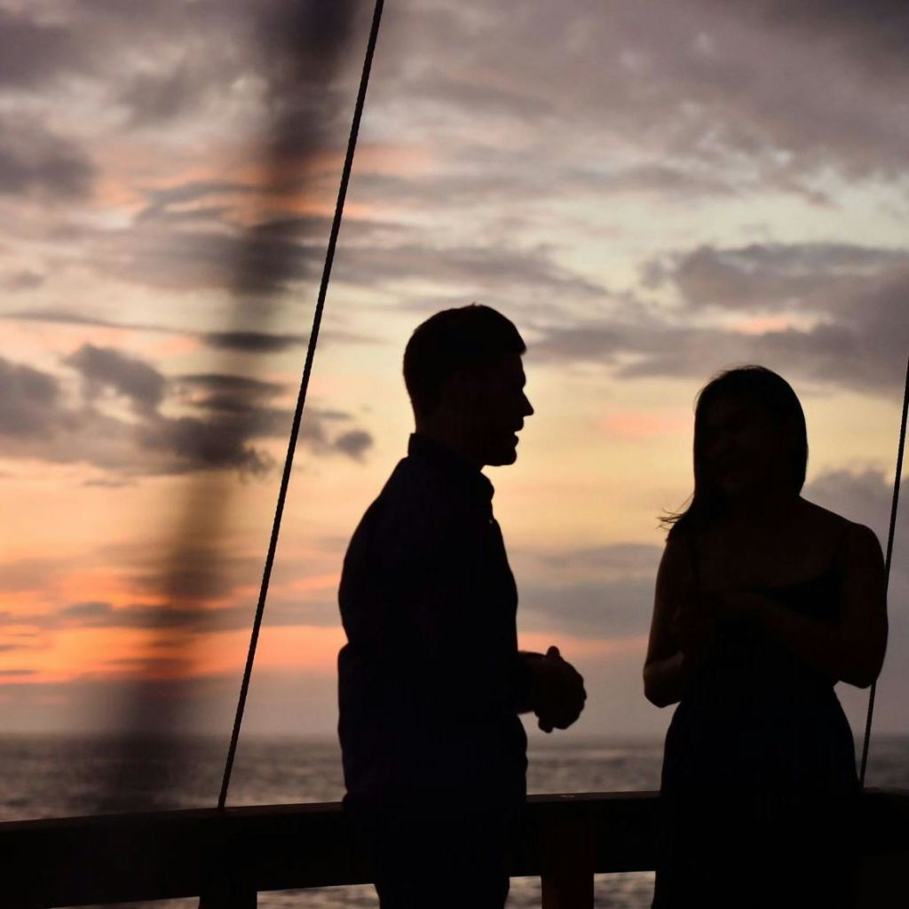 석양에 하늘과 바다를 배경으로 두 사람이 갑판에 서 있습니다. 로프 또는 케이블 라인이 부분적으로 보입니다.