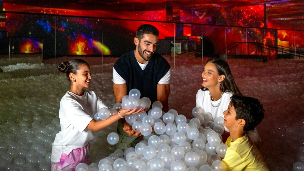 어른 두 명과 어린이 두 명이 반투명 공이 가득한 공 구덩이에서 화려한 조명 아래에서 웃으며 즐겁게 놀고 있습니다.