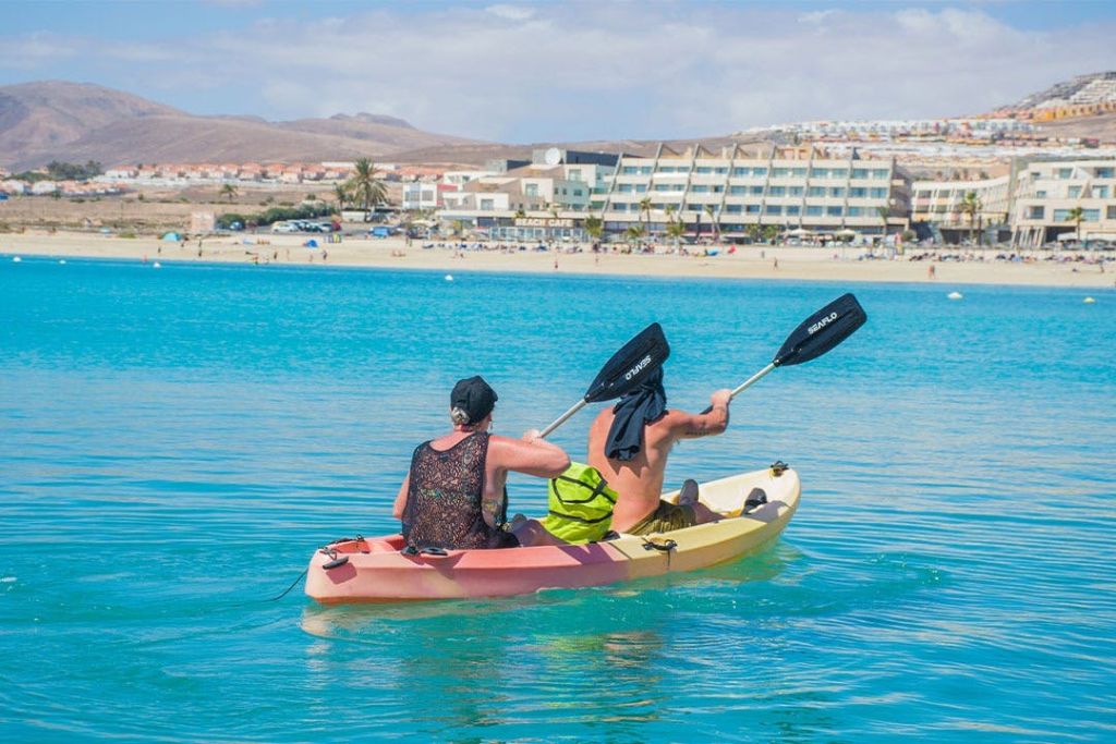 호텔과 산을 배경으로 모래 해변 근처의 맑고 푸른 바다에서 두 사람이 카약을 타고 노를 젓고 있습니다.