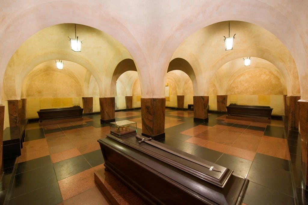 타일 바닥에 아치형 천장, 매달린 등불, 여러 개의 밀폐된 관이 놓인 지하 방.