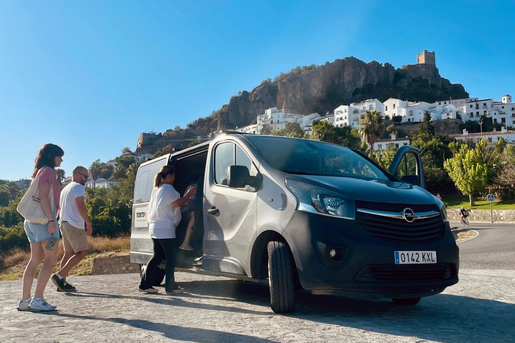한 사람이 하얀 건물과 바위 언덕을 배경으로 햇살이 내리쬐는 야외 공간에서 다른 사람을 검은색 밴으로 안내합니다.