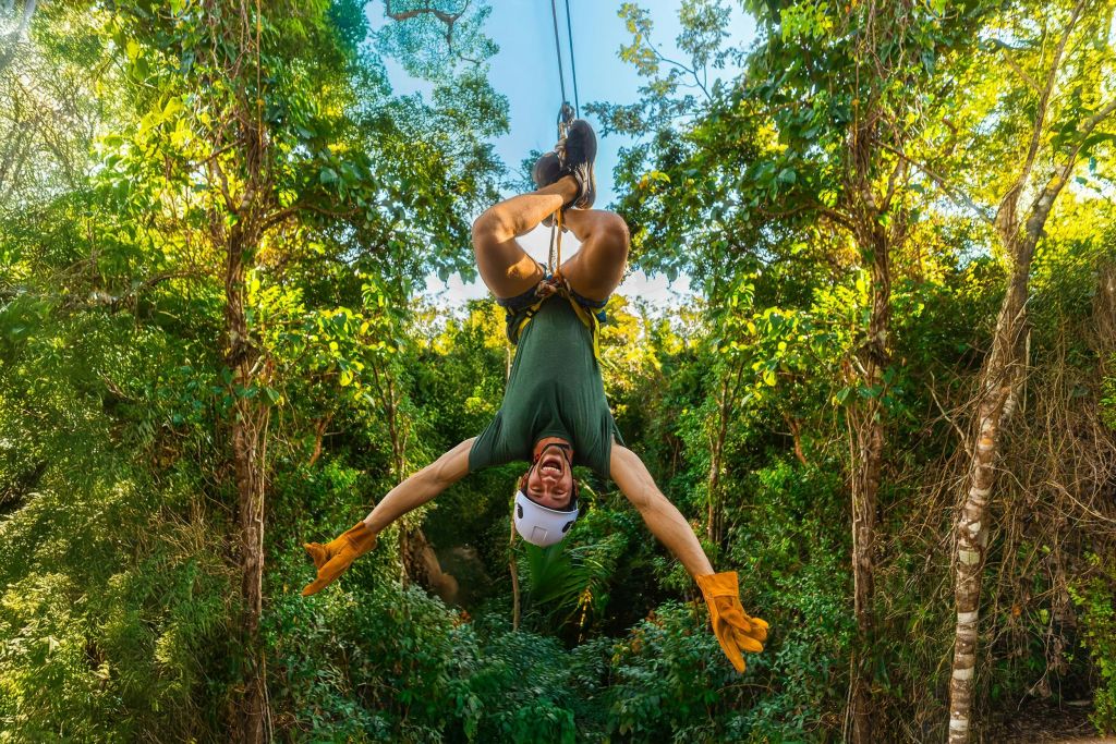 숲 속 짚라인에 거꾸로 매달려 있고, 무성한 녹음으로 둘러싸인 헬멧, 장갑, 하네스를 착용한 사람.