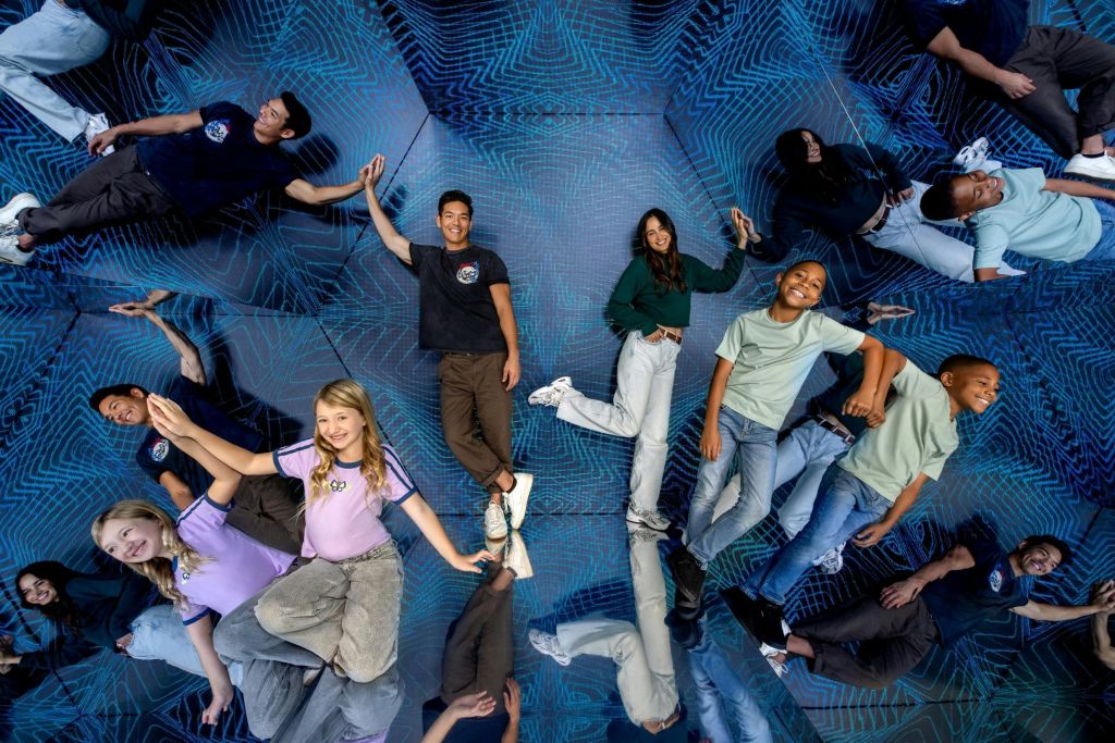 거울 벽과 파란색 기하학적 패턴이 있는 방에서 포즈를 취하는 네 명의 사람이 주변을 비추며 포즈를 취하고 있습니다.