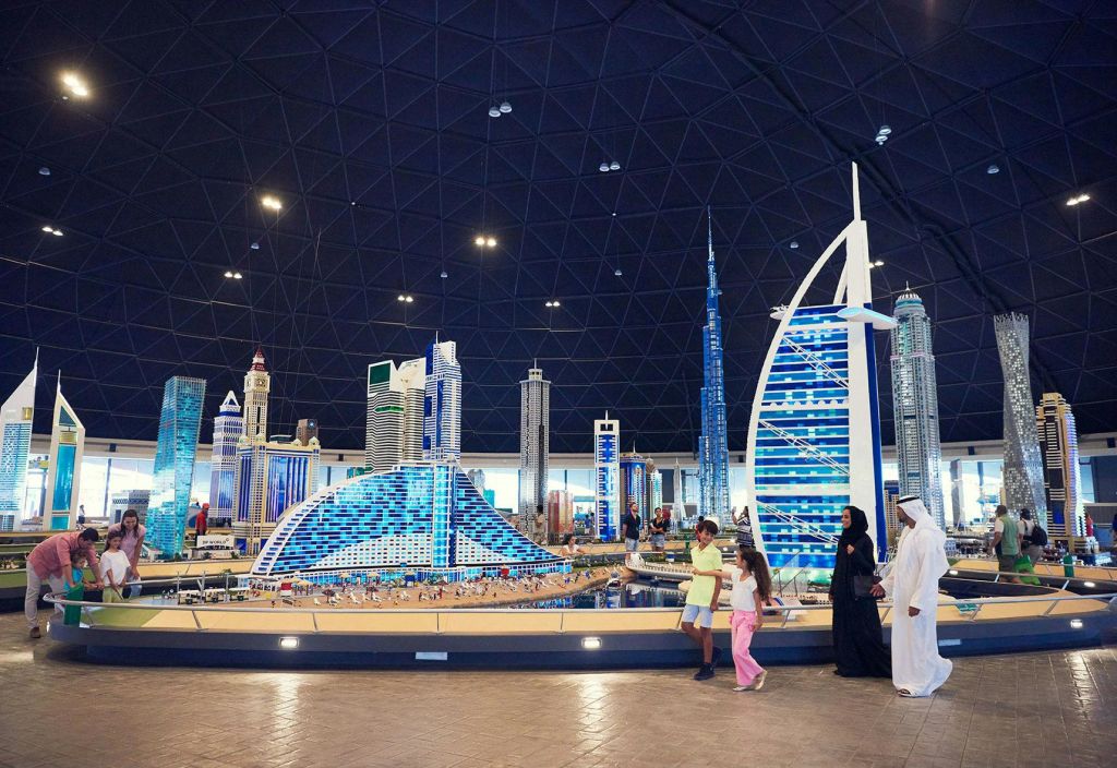 돔 구조물 내부에서 상징적인 고층 빌딩을 포함한 도시의 스카이라인을 자세히 보여주는 레고 모델을 따라 걷는 사람들.