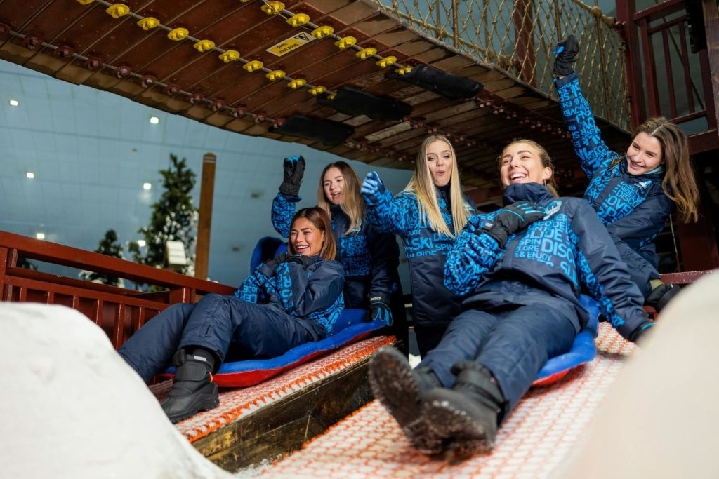 파란색과 검은색 겨울 장비를 착용한 네 명이 실내 스노우 슬로프에서 미끄러지듯 미소를 지으며 흥분한 듯 손을 들어 올립니다.