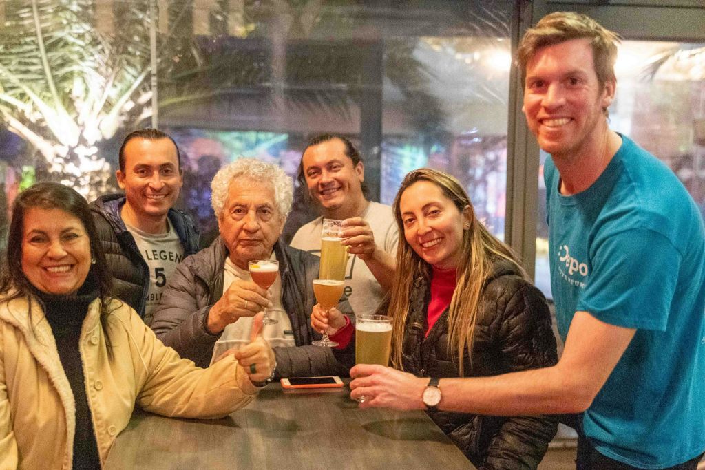 테이블에 둘러앉은 6명의 일행이 안경을 들고 미소를 짓고 있으며, 창문 너머로 축제 배경이 보입니다.