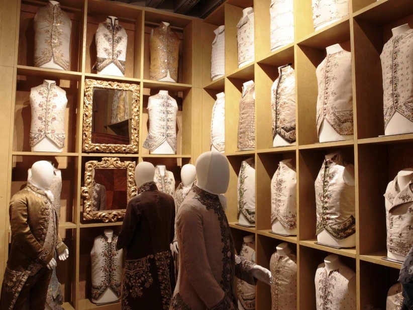 나무 입방체로 장식된 화려하고 유서 깊은 조끼와 비슷한 옷을 입은 마네킹, 벽에 장식용 거울이 전시되어 있습니다.