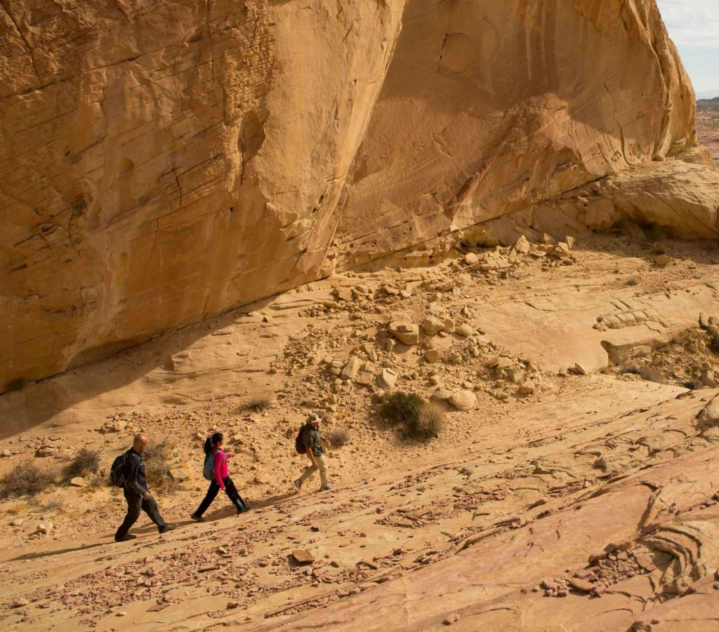 맑은 하늘 아래 커다란 사막 절벽의 바위 언덕을 따라 세 사람이 하이킹을 하고 있습니다.