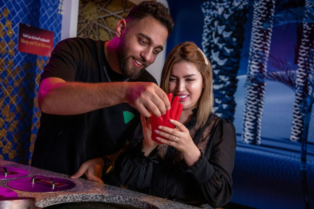 두 사람이 함께 미소를 짓고 붉은 왁스의 손을 성형합니다. 배경에는 추상적인 파란색과 흰색 패턴이 있습니다.