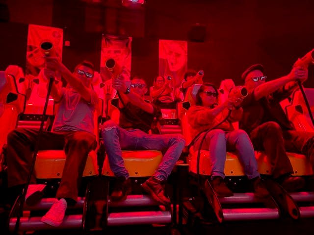 3D 안경을 착용한 사람들은 어두운 적색 조명 게임 환경에 푹 빠져 게임 컨트롤러를 들고 빨간색 좌석에 앉아 있습니다.