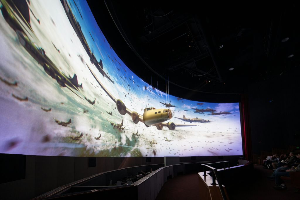 어두운 방의 곡선형 화면은 제2차 세계 대전 시대의 비행기가 지층을 이루어 풍경 위로 날아가는 디지털 투영을 보여줍니다.