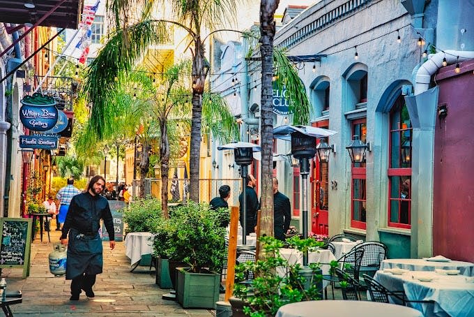 테이블, 의자, 화분, 야자수 아래 통로를 따라 걷는 사람들이 있는 활기찬 야외 식사 공간.