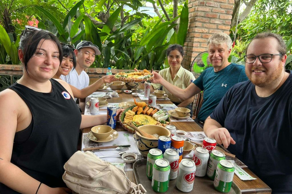 세 사람이 야외 테이블에 앉아 푸르른 녹음에 둘러싸인 다양한 음식과 음료를 즐기고 있습니다.