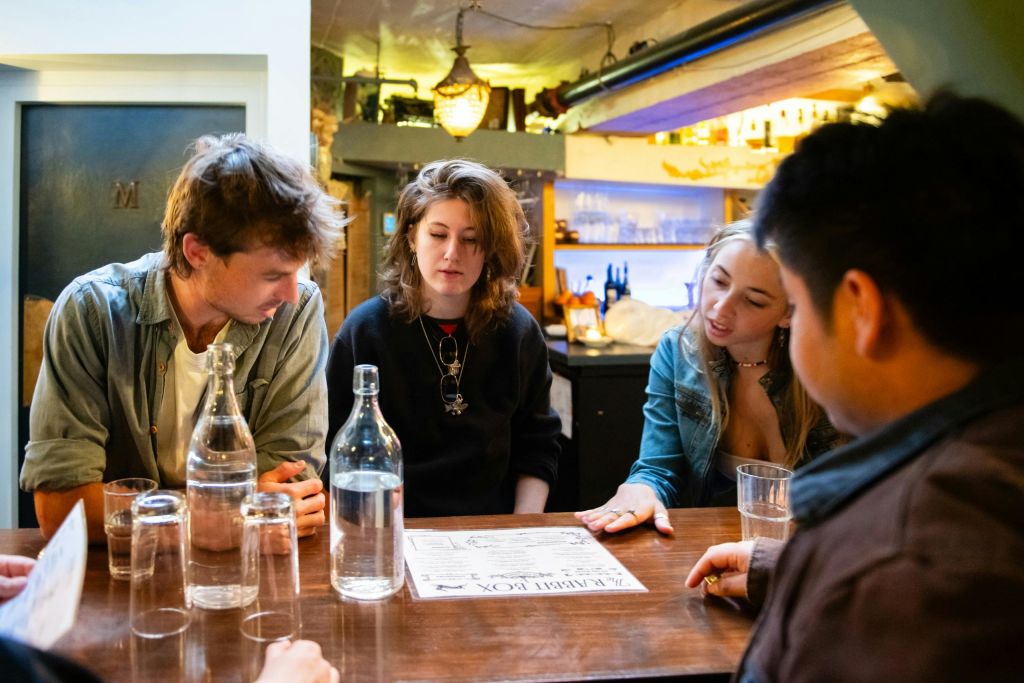 세 사람이 아늑한 인테리어의 나무 테이블에 앉아 테이블 위에 놓인 유리잔과 병이 놓인 메뉴에 대해 이야기를 나누고 있습니다.