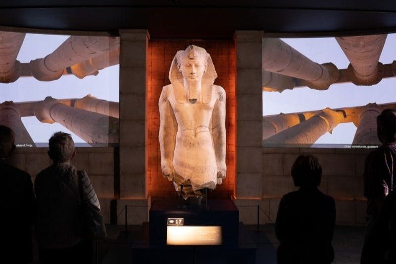 박물관에 전시된 대형 이집트 동상으로, 두 사람이 그것을 관찰하고 있으며, 사원 기둥을 배경으로 세워져 있습니다.