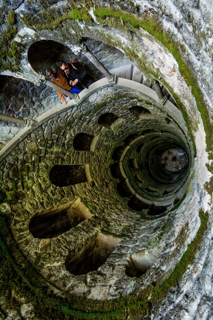 갈색 재킷을 입은 사람이 여러 개의 아치형 개구부와 이끼로 덮인 벽이 있는 돌 나선형 계단을 내려다보고 있습니다.