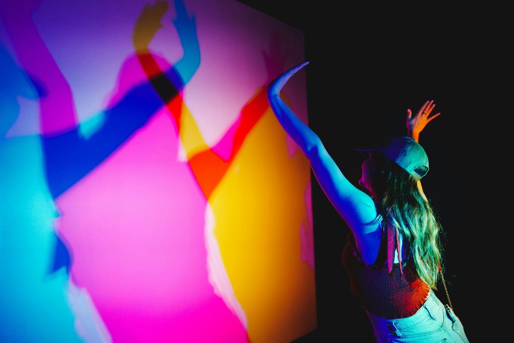 다색 조명 설치와 상호 작용하는 캡의 사람이 벽에 겹쳐진 그림자를 드리웁니다.