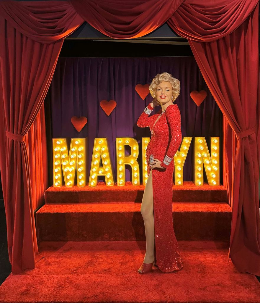 빨간색 무대를 배경으로 "마릴린" 글자와 하트 장식이 빛나는 레드 드레스를 입은 왁스 피규어가 포즈를 취하고 있습니다.