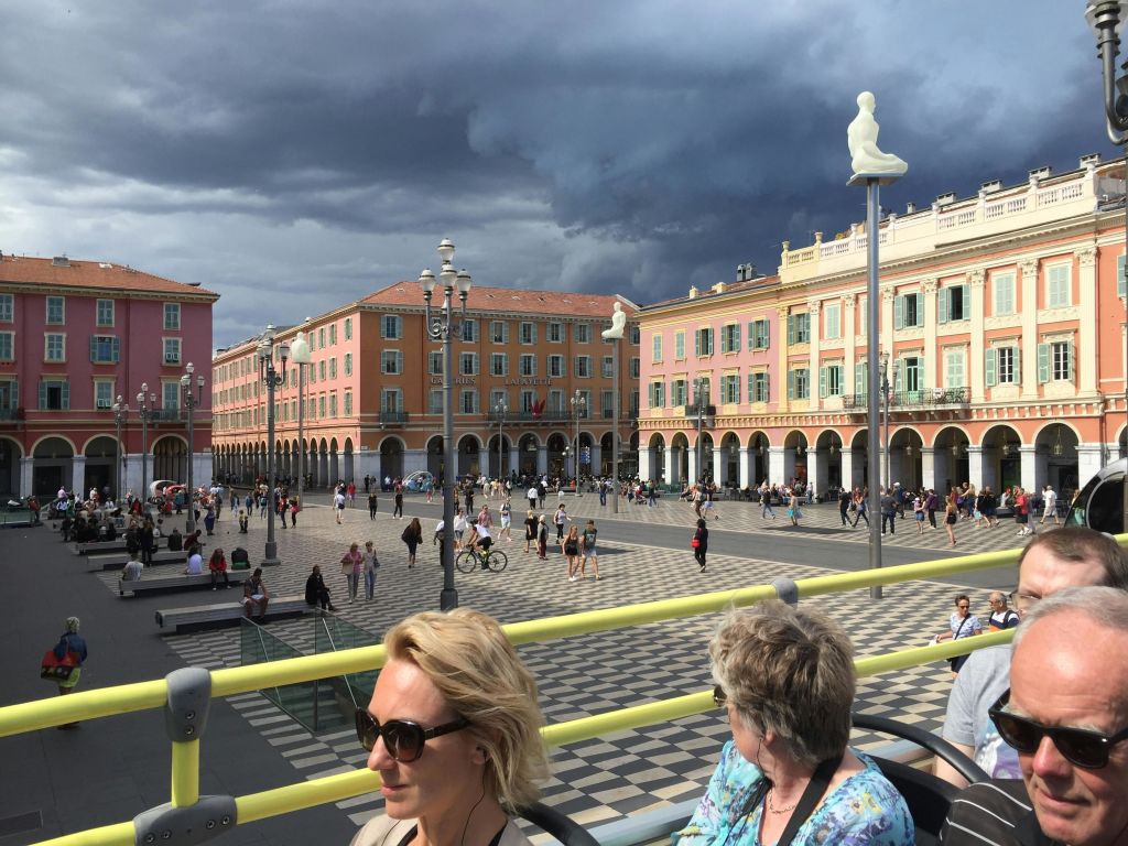 사람들은 흐린 하늘 아래 화려한 건물로 둘러싸인 넓은 광장에서 걷고 어울립니다. 두 명의 여성이 전경에 앉아 있습니다.