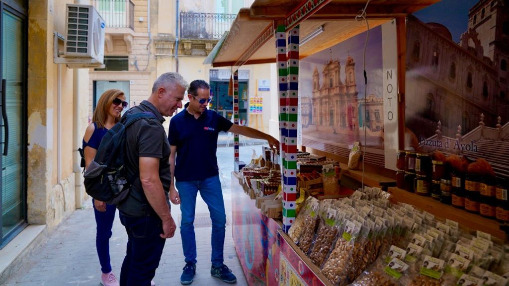 건물과 에어컨 장치가 있는 좁은 거리의 다채로운 물품이 전시된 야외 가판대에 세 사람이 서 있습니다.