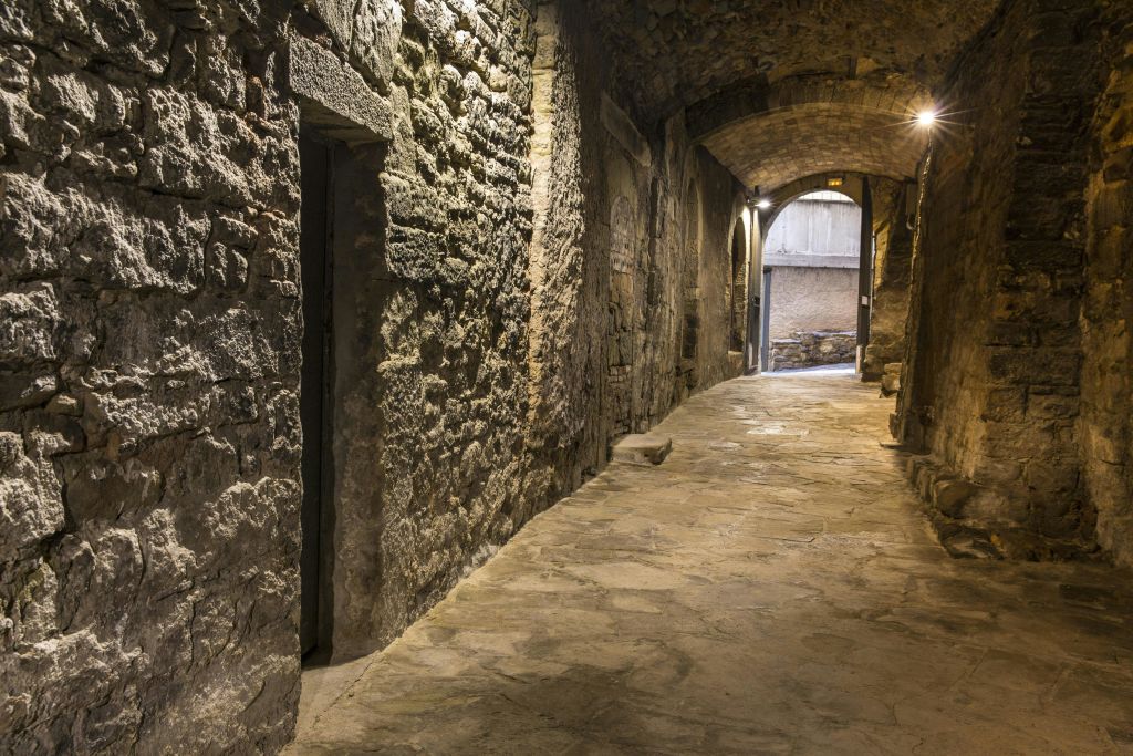 아치형 천장과 마모된 돌담이 있는 좁고 어두운 석조 통로로, 개방적이고 밝은 공간으로 이어집니다.