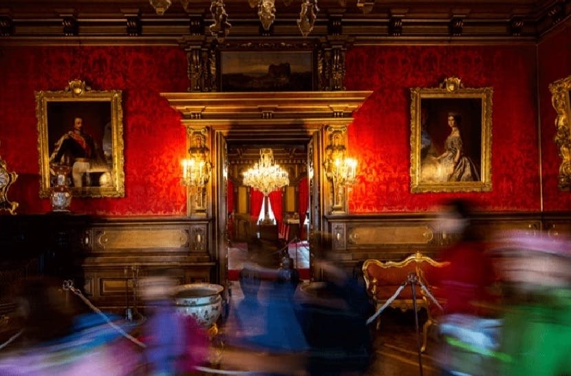 붉은 벽, 화려한 장식, 샹들리에, 초상화가 있는 호화로운 객실. 앞쪽에 흐릿한 형체가 무브먼트를 암시합니다.