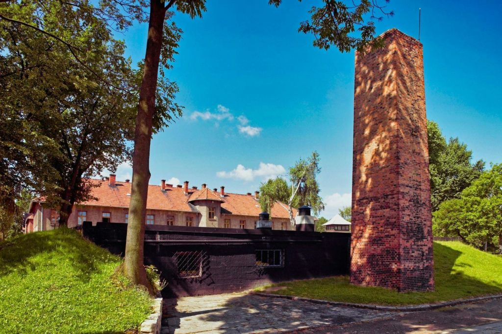 나무와 잔디로 둘러싸인 높은 굴뚝이 있는 벽돌 건물. 배경에는 맑고 푸른 하늘 아래 빨간 지붕의 건물이 보입니다.