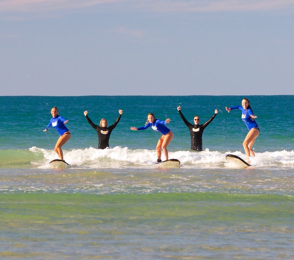 잠수복을 입은 다섯 명이 얕은 바다 파도에서 서핑을 하며 열정적으로 팔을 들어 올립니다.