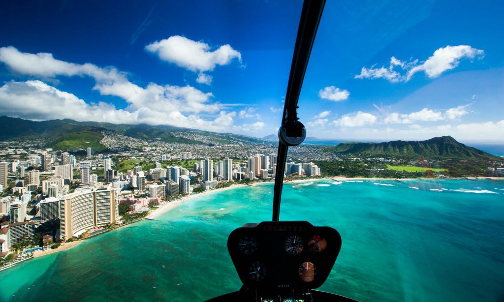 푸른 하늘 아래 청록색 바다, 하얀 모래 해변, 건물이 있는 해안 도시 위를 날아다니는 헬리콥터 조종석에서 바라보는 풍경.
