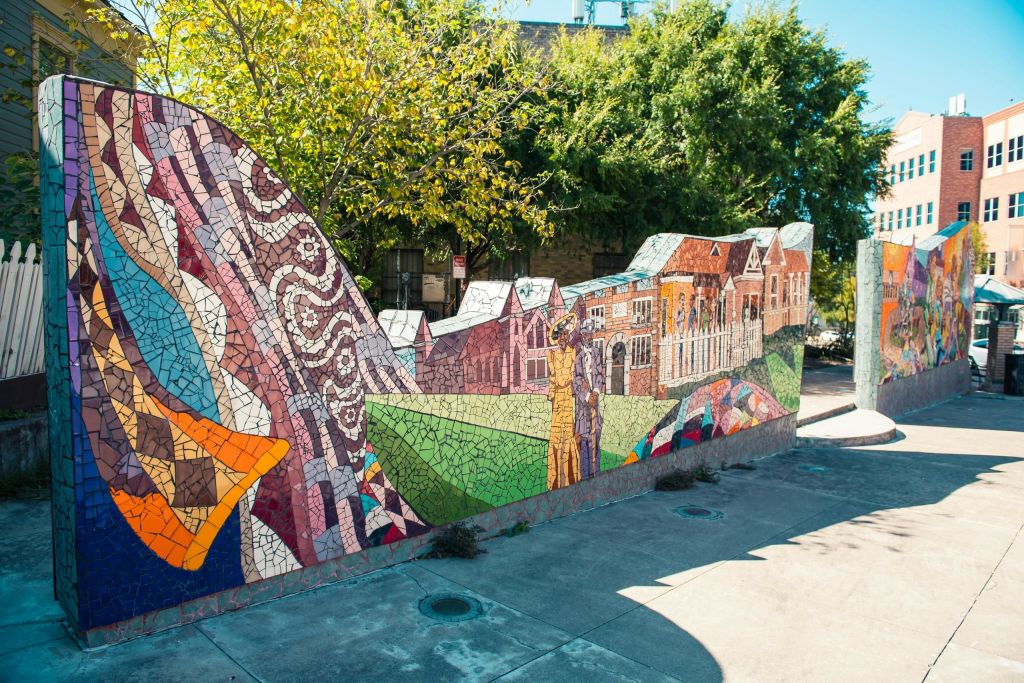 나무가 있는 야외 도시 환경을 배경으로 다채로운 무늬, 건물, 인물이 그려진 생동감 넘치는 모자이크 벽화입니다.