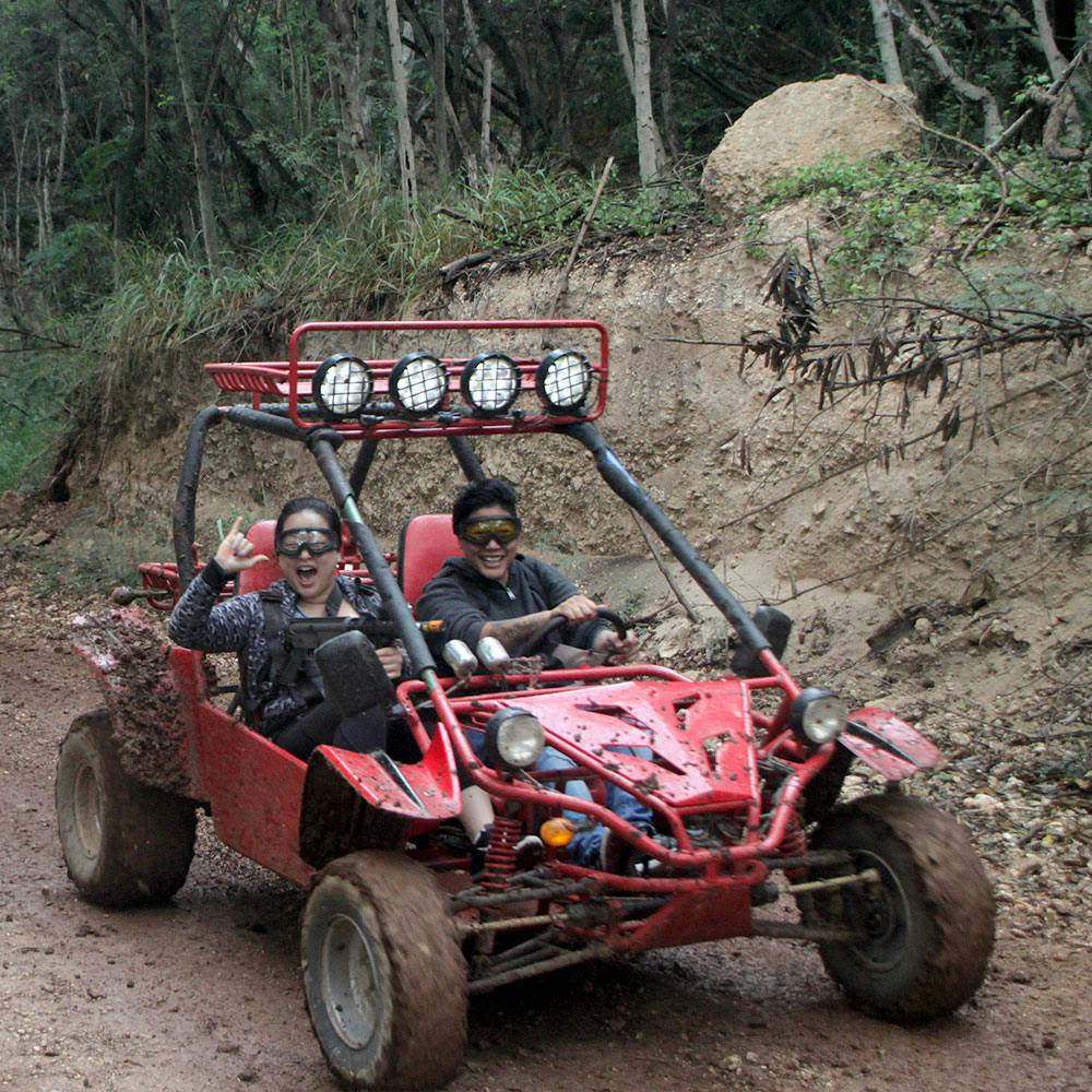 나무와 흙으로 둘러싸인 진흙 투성이 산책로에서 빨간색 오프로드 버기를 운전하는 두 사람. 한 사람이 손을 흔들고 있으며 둘 다 고글을 착용합니다.