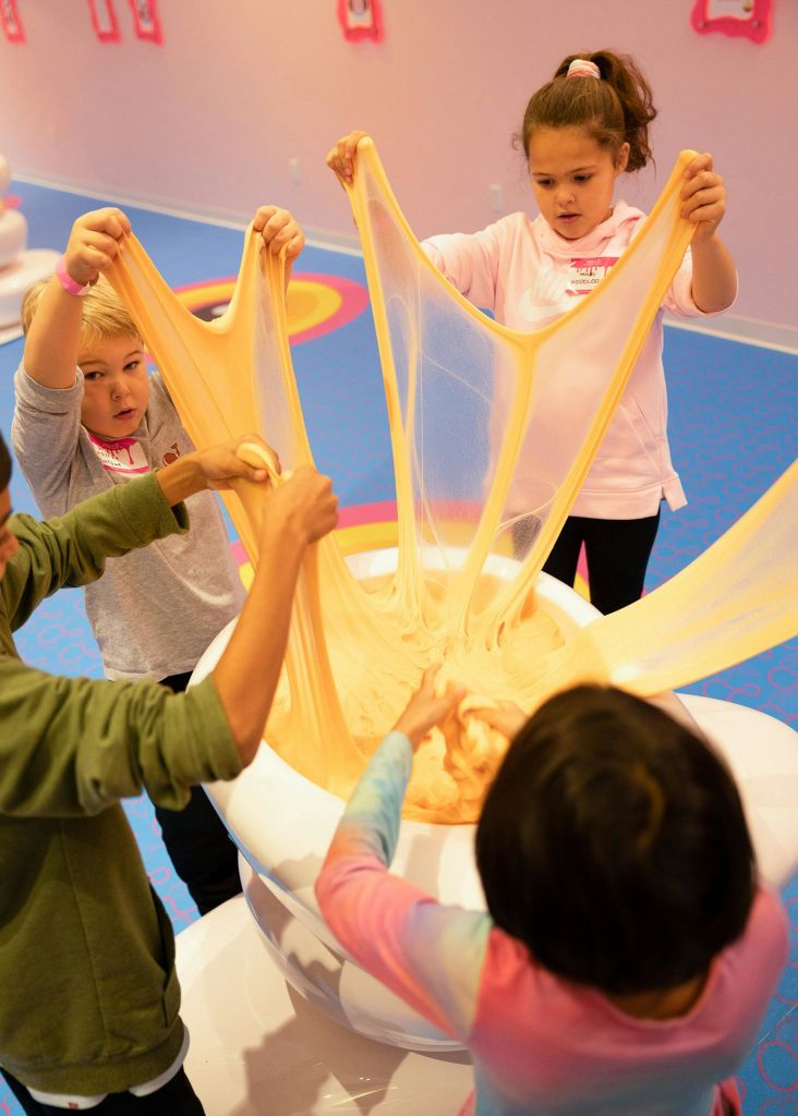 분홍색 장식과 파란색 바닥이 있는 놀이 센터에서 네 명의 어린이가 커다란 슬라임을 뻗어 있습니다.