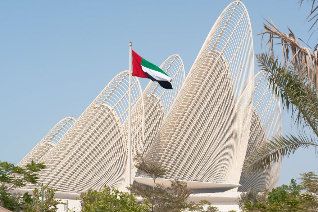 기둥에 달린 아랍에미리트 국기로, 맑은 하늘 아래를 배경으로 모던하고 곡선된 건축 구조물이 있습니다.