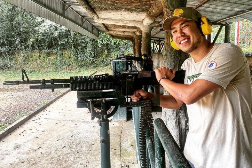 귀를 보호하고 모자를 쓴 남성이 야외 사격장에서 커다란 총을 조종하며 미소를 짓고 있습니다.