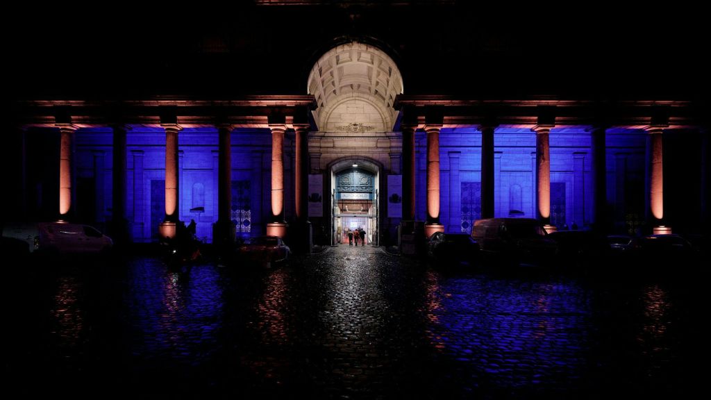 파란색과 주황색 조명, 기둥, 자갈길이 있는 야간 조명 건물 입구입니다. 멀리서 사람들이 들어온다.