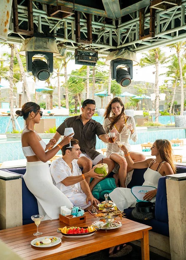 수영장과 야자수를 배경으로 야외 라운지에서 음료와 음식을 즐기는 흰색과 베이지색 의상을 입은 친구들.