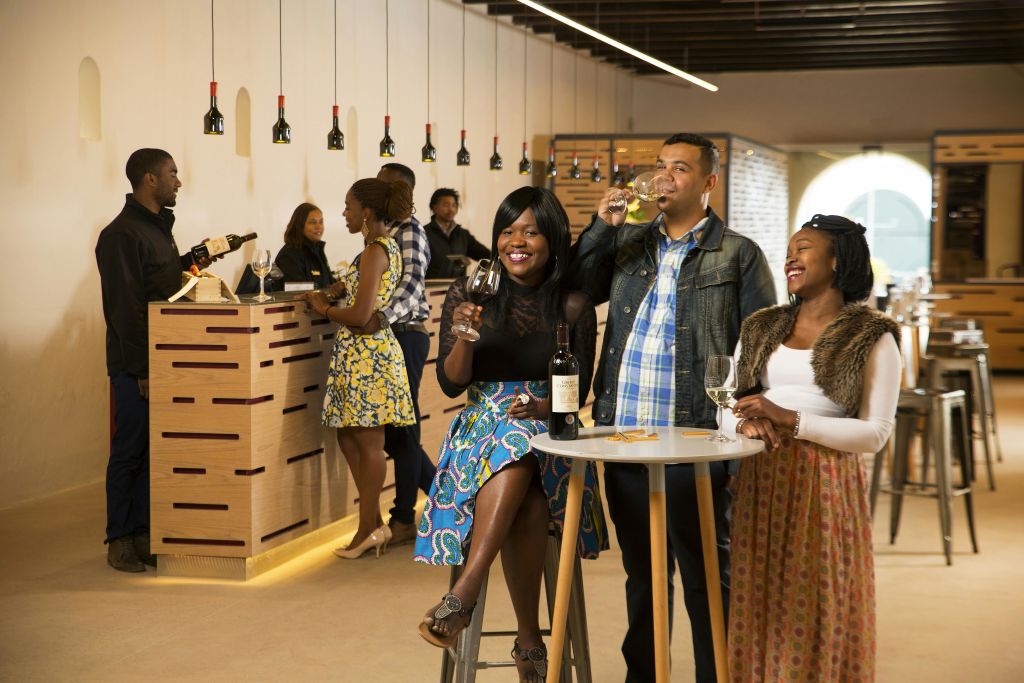 조명이 밝은 실내 와인 바에서 와인을 즐기고, 병과 잔이 놓인 작은 테이블 주변에 서 있는 사람들.