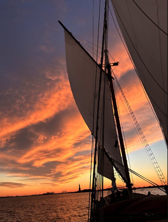 석양에 큰 돛을 단 범선으로, 저 멀리 활기찬 주황색 하늘 아래 자유의 여신상이 보입니다.