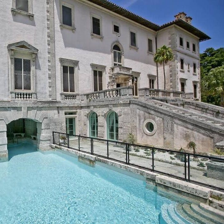 야자수와 녹지로 둘러싸인 청록색 수영장으로 이어지는 석조 계단이 있는 역사적인 대형 저택입니다.