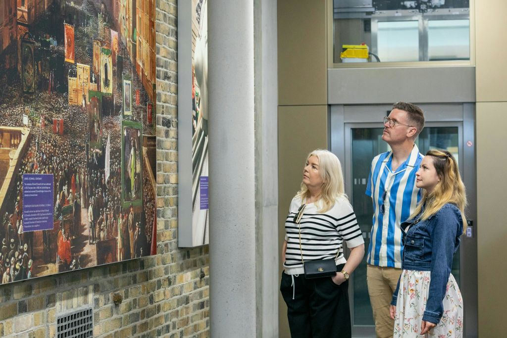 현대적인 실내 공간에서 세 사람이 벽을 관찰하며 엘리베이터와 벽돌 벽 근처에 서 있습니다.