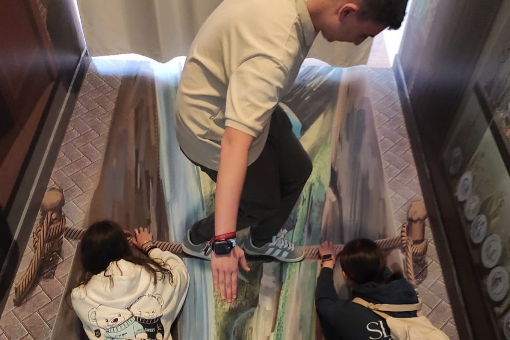 세 사람이 협곡 위의 밧줄 다리의 바닥 그림과 상호 작용하여 3D 착시 효과를 만듭니다.