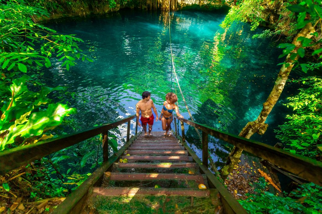 수영복을 입은 두 사람이 푸르른 단풍으로 둘러싸인 투명한 청록색 천연 수영장에서 나무 계단을 오르고 있습니다.